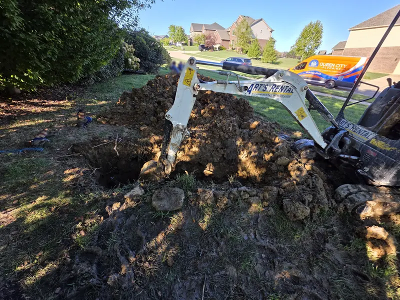 Mini excavator digging a trench for a main line repair, with a Queen City Plumbing van in the background