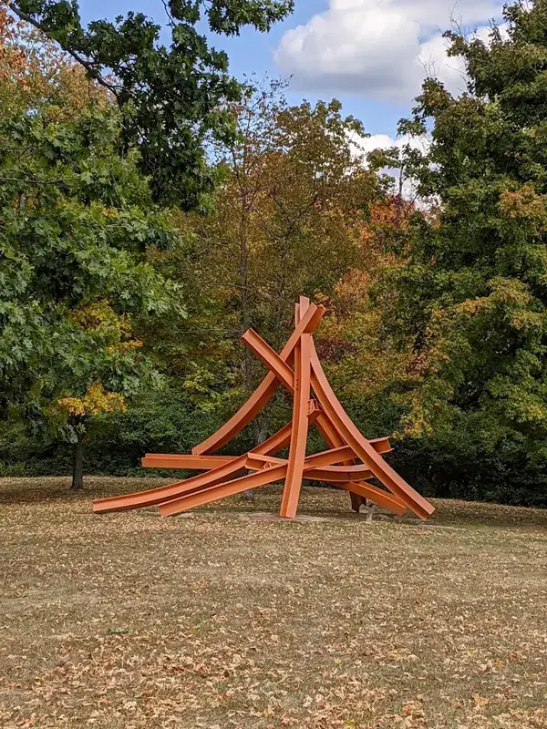 Orange steel beam sculpture on a lawn among autumn trees at Pyramid Hill in Hamilton