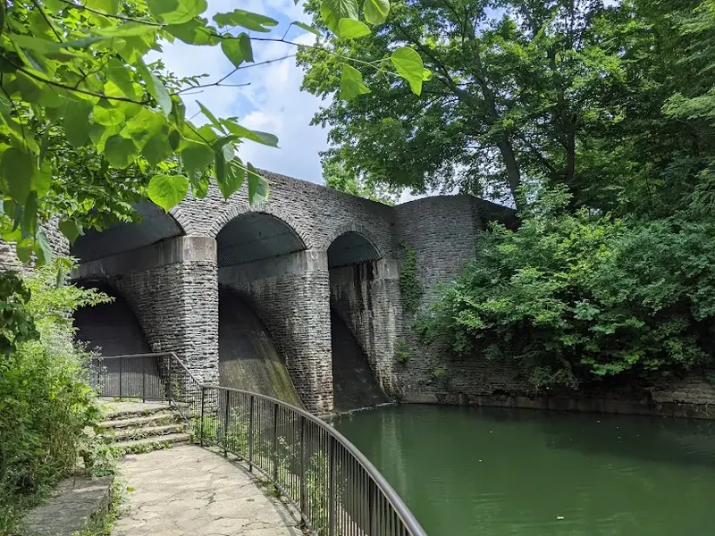 Stone dam with large arches over green water and a railed path at Sharon Woods