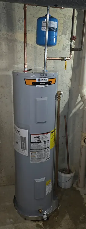 New State ProLine electric tank water heater with a blue expansion tank and copper piping against a concrete basement wall