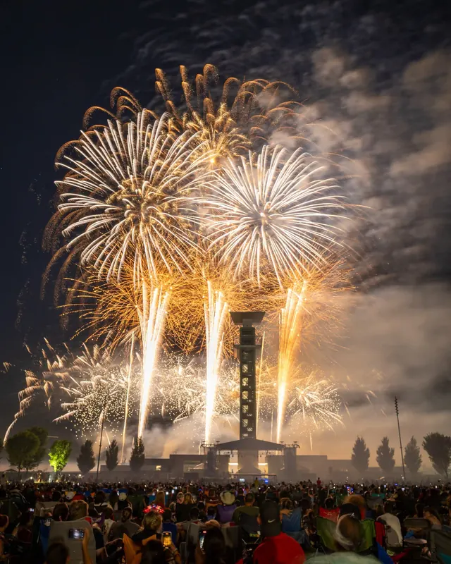 Fireworks bursting over the Summit Park observation tower with a crowd watching on the lawn