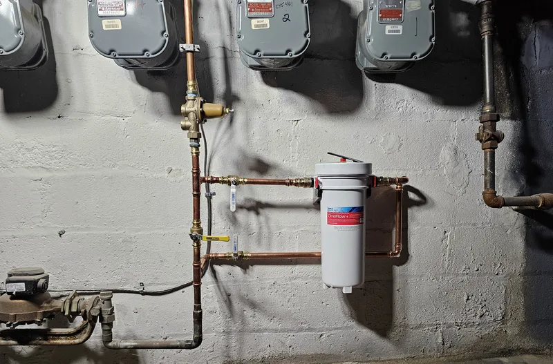 White whole-house water filter plumbed into copper piping on a basement wall beside gas meters
