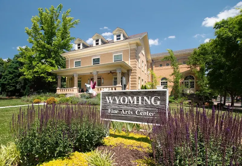 Wyoming Fine Arts Center, a yellow brick house with its sign and purple flowers out front